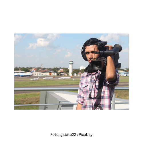 Ein Reporter mit einer Kamera auf der Schulter steht auf einem Flugplatz am Land.