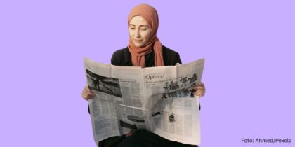 Das Bild zeigt eine junge Frau mit Kopftuch. Sie liest eine Printzeitung. Sie lächelt.